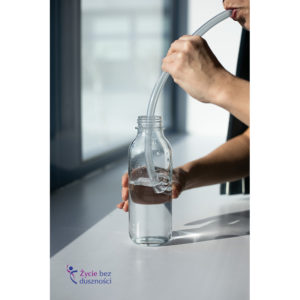 Zdjęcie obrazujące ćwiczenie oddechowe LAX VOX, z dmuchaniem przez rurkę o średnicy 1 cm do półlitrowej szklanej butelki wypełnionej wodą